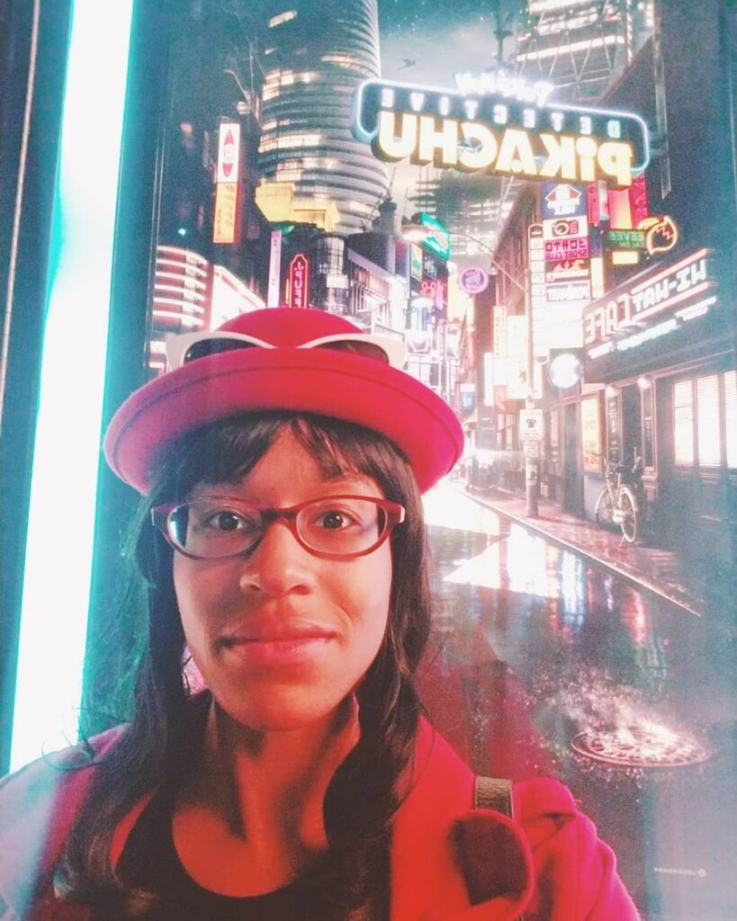 Me dressed like Serena from Pokémon taking a selfie in front of the Detective Pikachu movie poster